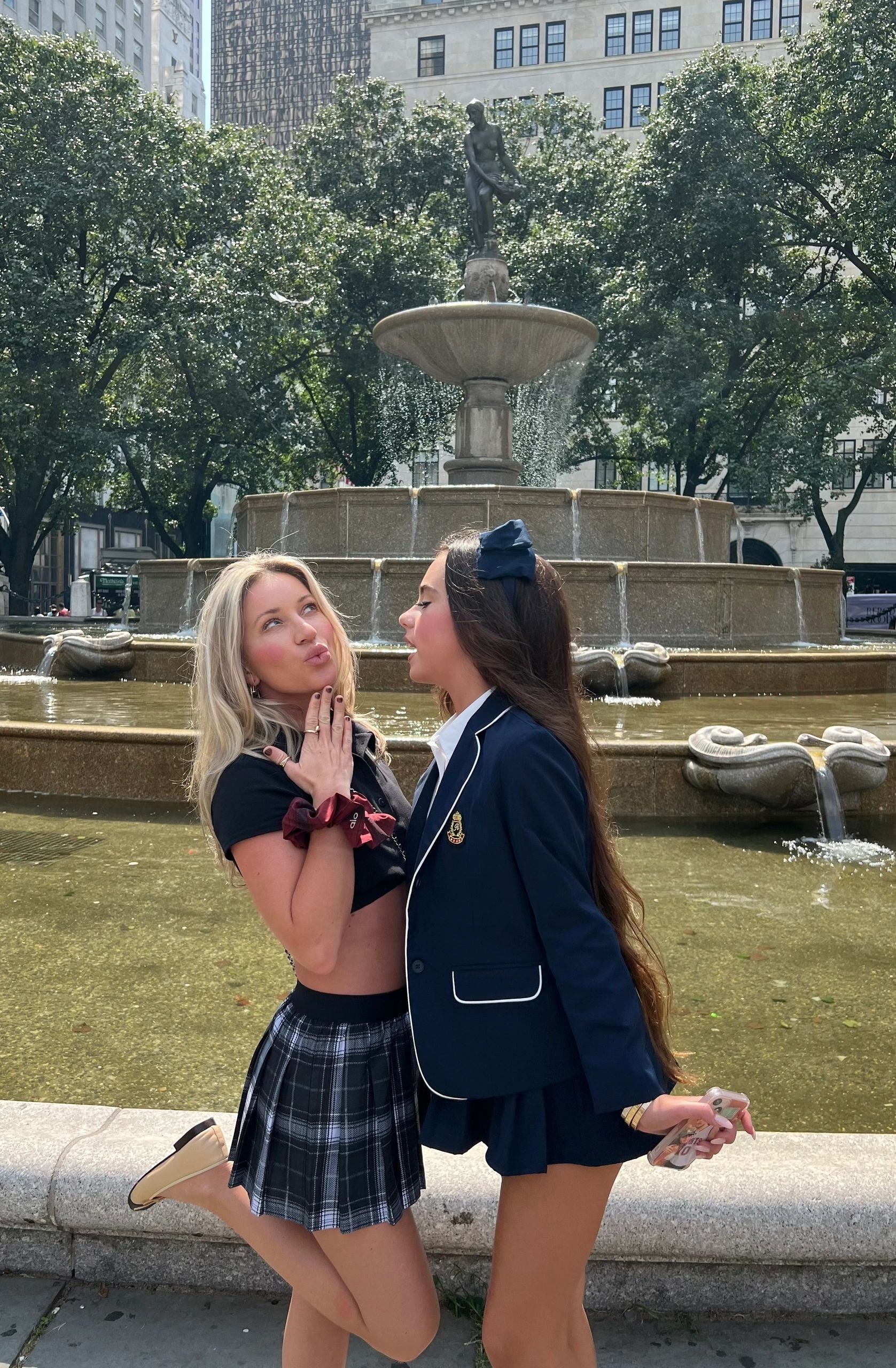 Two Gossip Girl Tours guests in plaid and prep-school outfits at the Pulitzer Fountain before the Bus and SUV tour in NYC